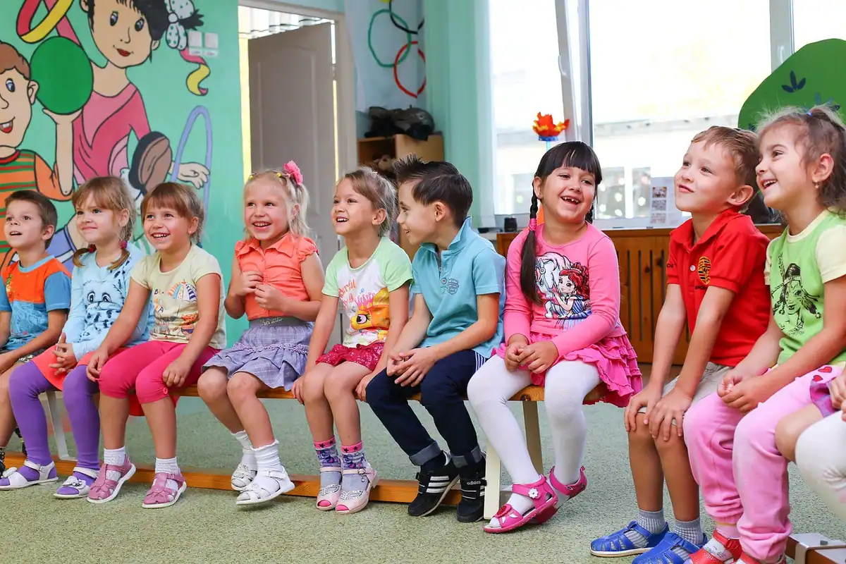 Grup de copii veseli, așezați pe o bancă într-o sală de grădiniță colorată