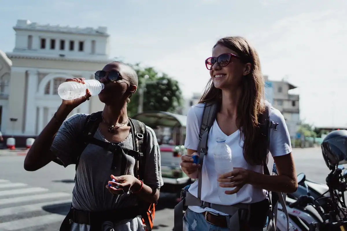 Doi turiști cu rucsaci care beau apă îmbuteliată în timp ce explorează un oraș