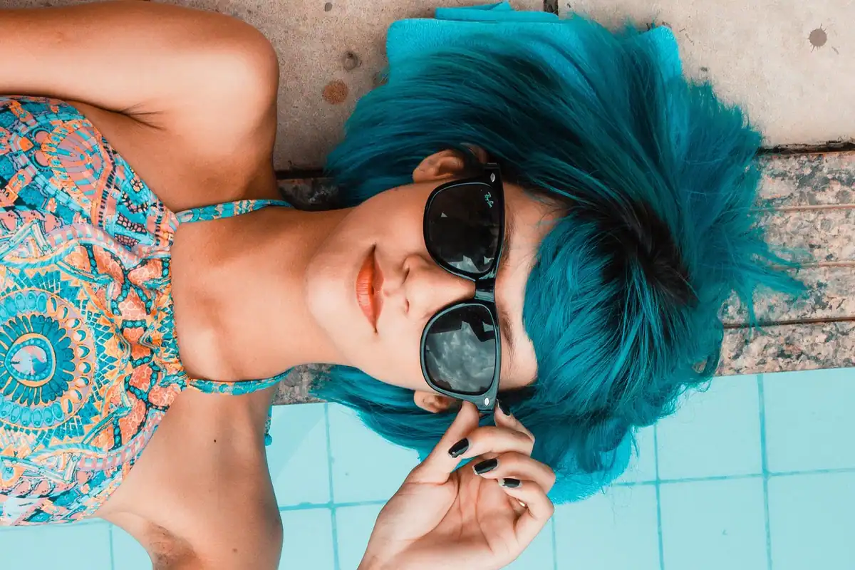Femeie cu păr albastru și ochelari de soare întinsă lângă o piscină