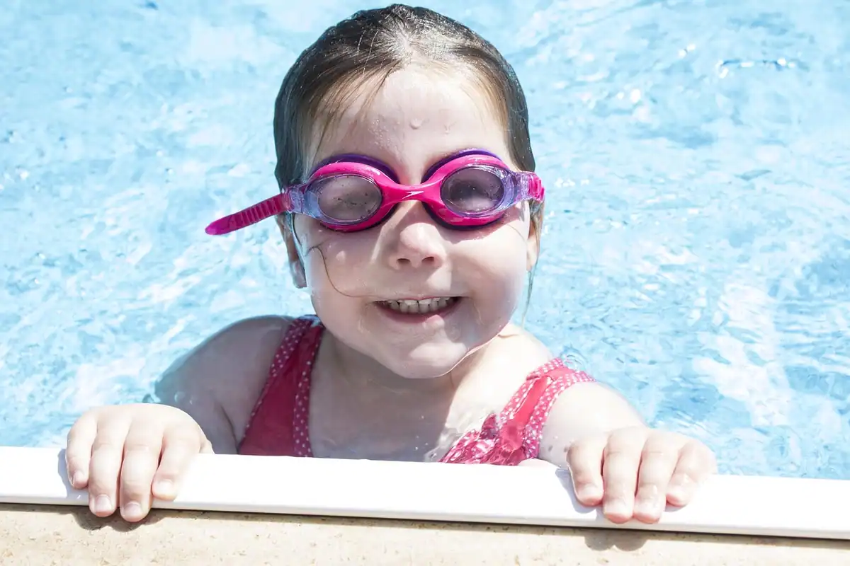 Fetiță zâmbitoare cu ochelari de înot roz, care se sprijină de marginea piscinei