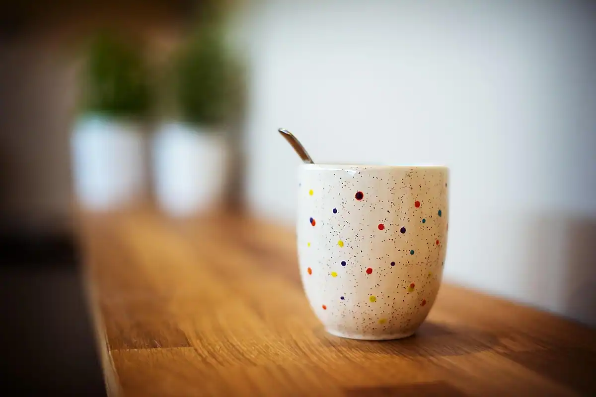 Cană ceramică cu buline colorate, cu o linguriță, așezată pe o masă din lemn
