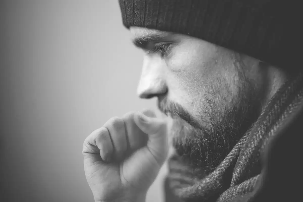 Bărbat cu căciulă și barbă, în alb-negru, care tușește cu pumnul la gură