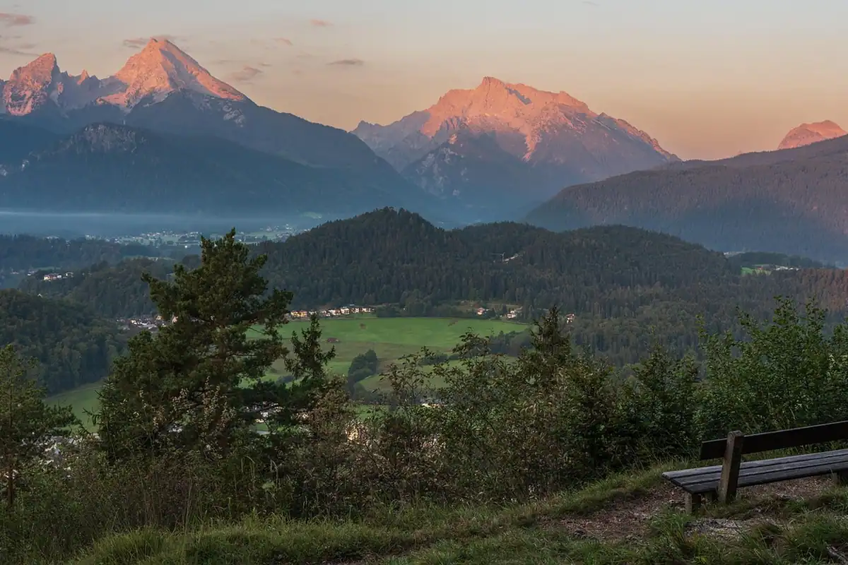 Munți cu vârfuri roz la răsărit, cu pădure, bancă și ceață pe vale
